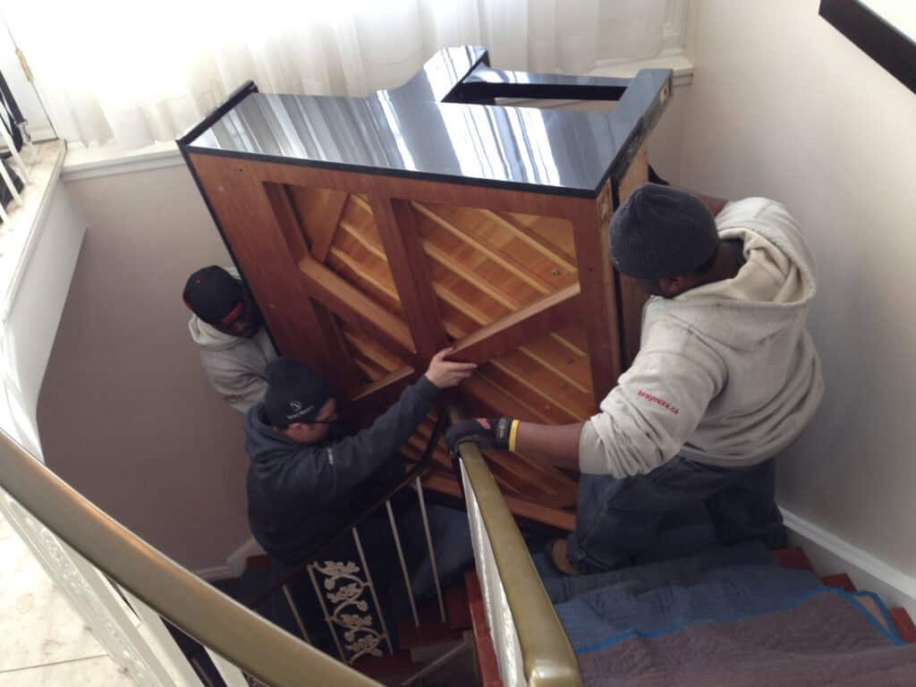 piano movers taking an upright piano down a flight of stairs to basement