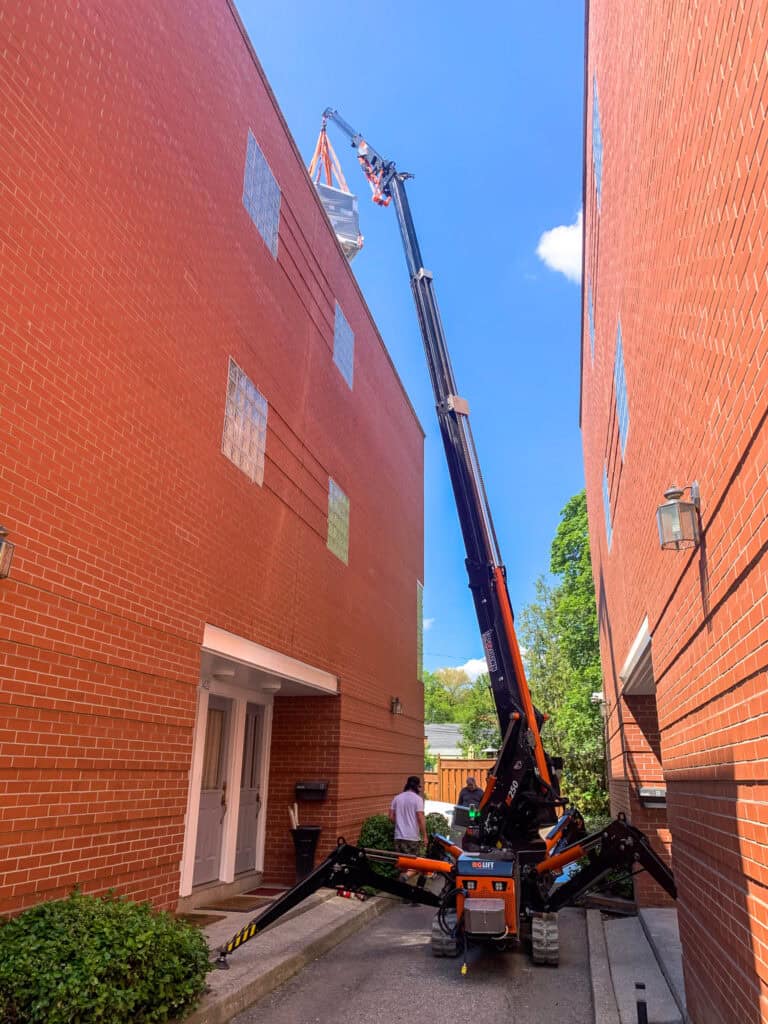 Spider crane in a tight alleyway between buildings, lifting an air conditioning unit up onto a roof.