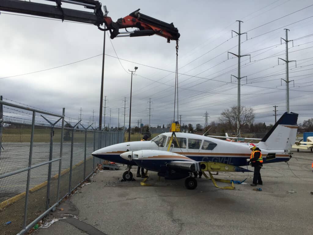 Knuckle crane lifting a small air craft over a fence.