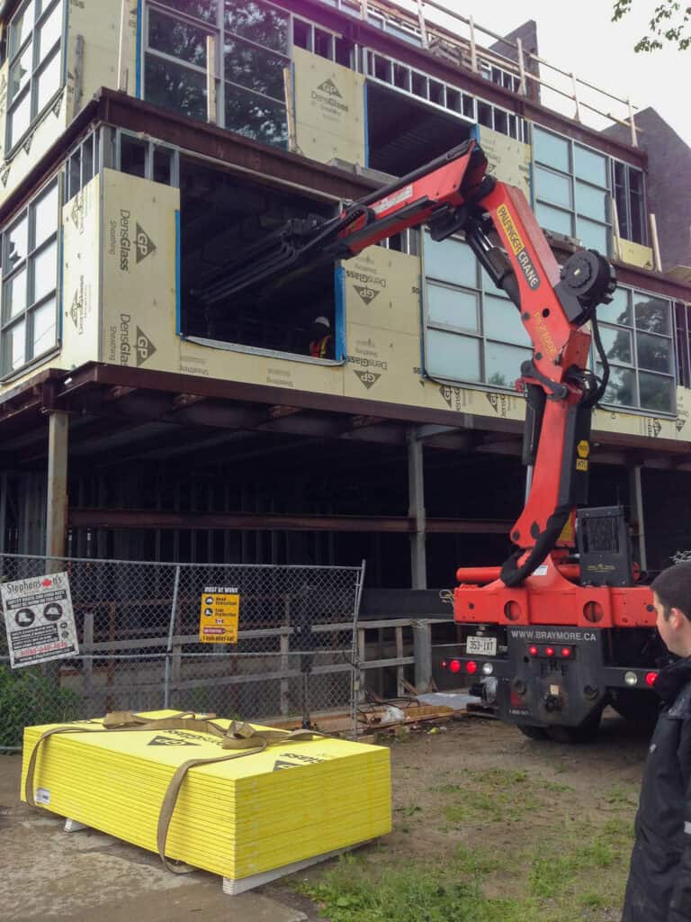 Knuckle crane lifting skids of plywood into building.