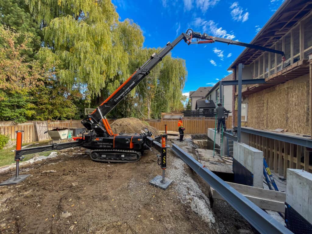 Mini crane in backyard being used to lift large steel beams in construction of a backyard addition to a house.