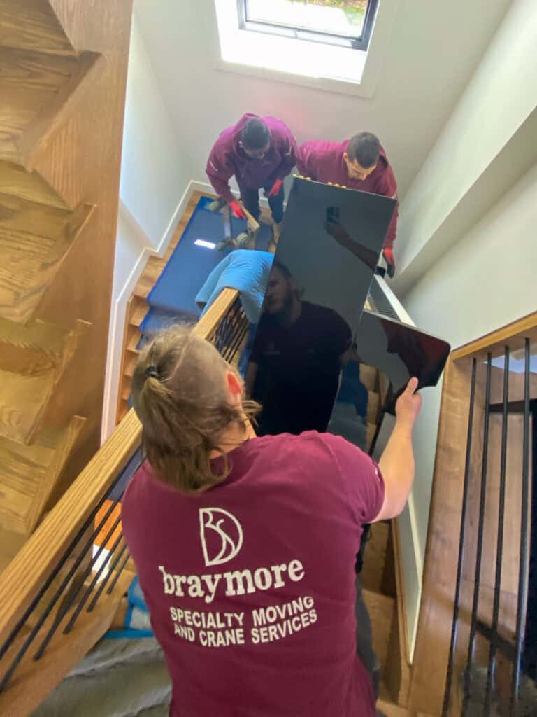Braymore Piano Movers are lifting an upright piano up a switch back staircase.